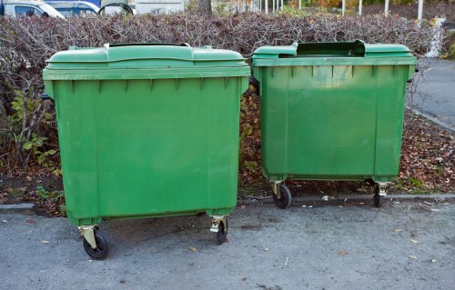 Two large green wheeled rubbish bins positioned side by side on a paved outdoor surface in a residential or commercial area with a background of shrubs and a fence. The bins are made of durable plastic with a corrugated texture and have fitted lids, with one bin slightly smaller and placed to the right of the larger one. They appear to be used for general waste disposal and are situated on a flat, tarmac-like pavement. The scene is well-lit, suggesting daytime, and the bins may belong to a waste collection service such as Commercial Waste Docklands serving the Docklands area, with the postcode context subtly implied by the surroundings. The overall setting indicates a typical waste management setup for local rubbish removal, with the bins positioned for easy accessibility for waste collection or disposal purposes.