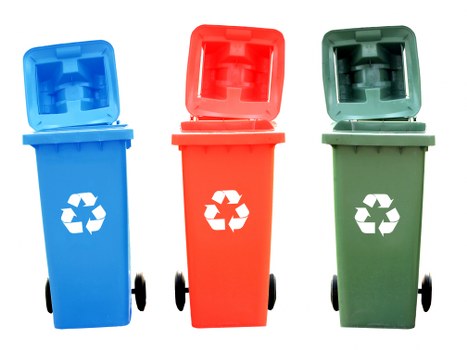 Three wheelie bins are positioned side by side on a paved surface, each with a distinct colour: blue, red, and green. The blue bin is on the left, featuring a secure open lid with a smooth, slightly textured plastic finish and a white recycling symbol on the front. The red bin is in the centre, with its lid partly open, showing a ribbed interior surface consistent with typical waste disposal bins, and also bears a white recycling symbol. The green bin stands on the right, with its lid fully open, revealing an interior with a slightly rough texture, suggesting suitability for compost or organic waste. Each bin is mounted on black wheels, which are positioned on the pavement, indicating they can be moved for waste collection. The background shows part of a building and a clear sky, suggesting an outdoor domestic or commercial setting, likely in an urban area of Docklands. The image is well-lit, with natural light highlighting the different textures and colours of the bins, consistent with a typical rubbish removal setup by Commercial Waste Docklands managing waste in the local area.