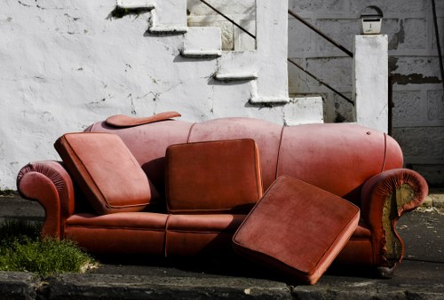 A worn, red-brown sofa with rolled arms and decorative wooden detailing on the armrests, positioned outdoors against a concrete wall with visible cracks and peeling paint. The sofa is partially collapsed, with some cushions displaced and a matching ottoman leaning against it, on a rough, asphalt surface with patches of green grass growing nearby. Behind the sofa, a set of concrete stairs with a metal railing is visible, indicating an urban setting likely near a residential or commercial building in Docklands. The lighting suggests daytime with natural sunlight illuminating the scene, highlighting the textures of the fabric and the weathered appearance of the furniture and wall. This image exemplifies the kind of furniture waste that Commercial Waste Docklands might handle as part of rubbish removal services in the area, combining elements of urban environmental neglect and furniture disposal needs.
