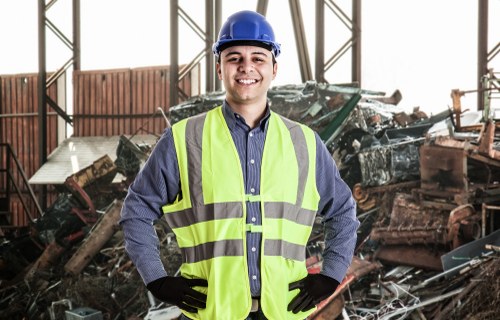A male waste management worker inside a warehouse or industrial facility, wearing a blue safety helmet, high-visibility yellow vest, and black gloves, standing in front of a large pile of mixed construction and industrial waste materials on the floor. The waste includes broken wood, metal scraps, and other debris with varying textures and colors, predominantly dark and rusty tones. The setting features metal framework and windows that allow natural light to illuminate the scene. The worker is smiling and has a confident posture, reflecting professionalism associated with rubbish removal services, similar to those provided by Commercial Waste Docklands near London. The environment suggests an active waste sorting or removal operation typical of the Docklands area in East London, supporting local rubbish management services.