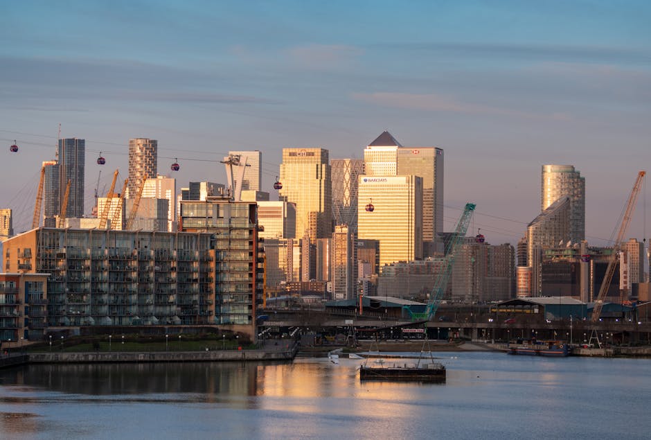 A wide-angle view of a modern cityscape during twilight, featuring tall commercial office buildings with glass facades reflecting ambient light and illuminated from within, situated along a waterway. 