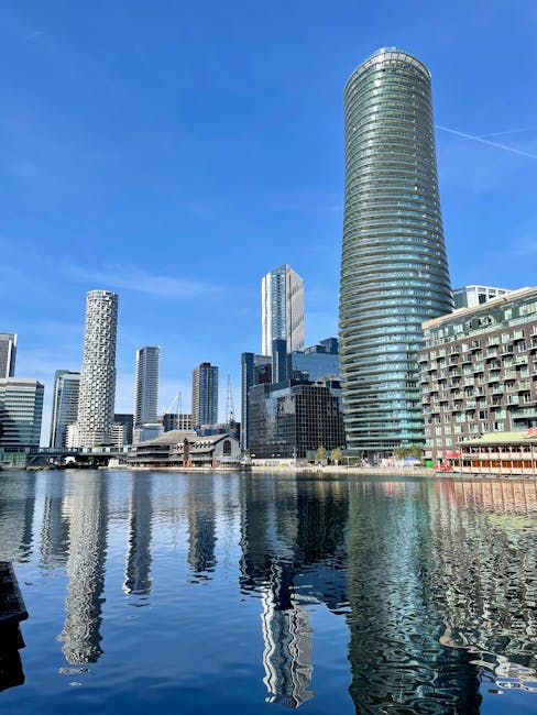 A collection of modern high-rise buildings situated along a waterfront, with reflective glass facades showing clear mirrors of the skyline in the calm water below. The tallest building on the right is